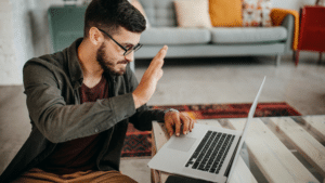 Photo of a man waving to his computer while working in a hybrid work enviroment 